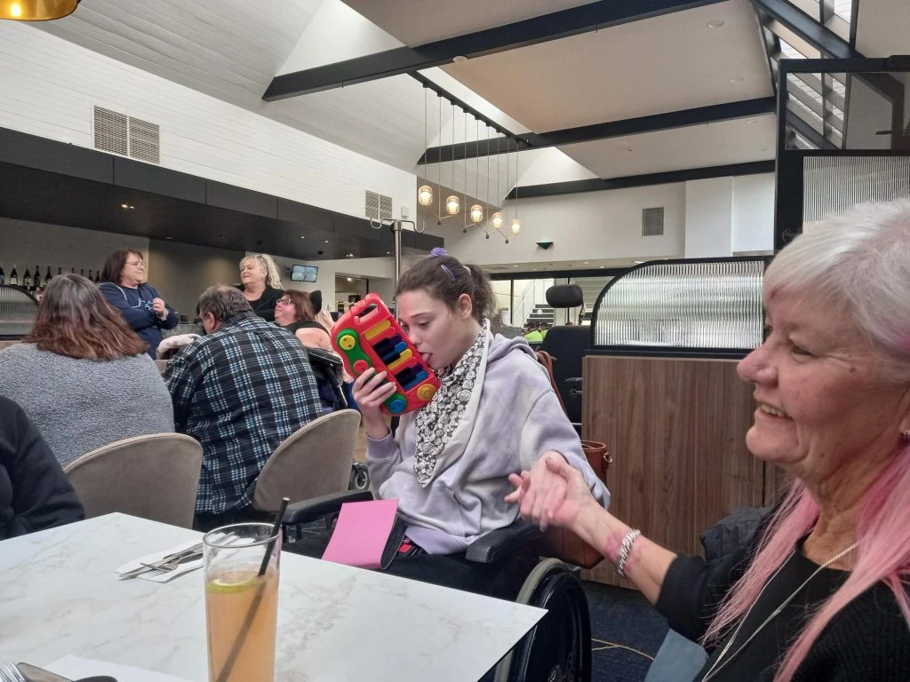 A young person sits in a wheelchair at a dining table, holding a colorful toy truck to their face, while another person with long white and pink hair sits nearby smiling. Other diners are in the background.