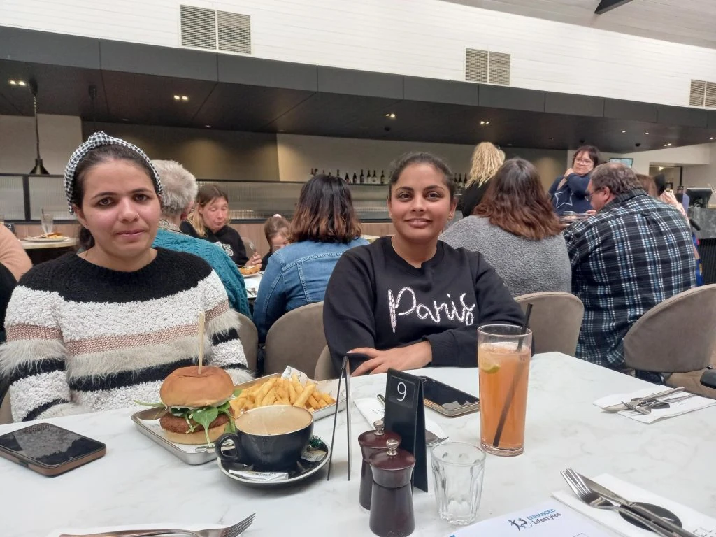 Two women sitting at a restaurant table. One has a burger and fries in front of her; the other has a drink. Other patrons are seated in the background.