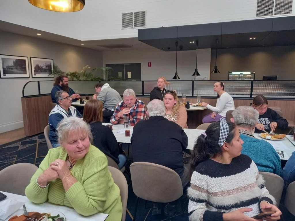 A group of people are seated at tables in a dining area, eating and conversing. The room has modern decor with light fixtures and artwork on the walls.