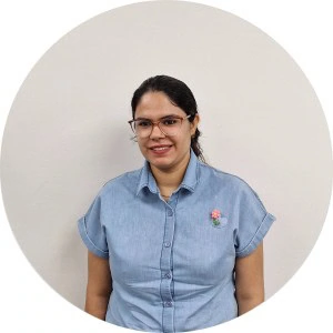 A person wearing glasses and a blue denim shirt with a flower brooch stands against a plain white background, smiling.