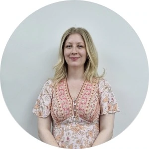 Person with long blond hair wearing a floral dress stands against a plain white background.