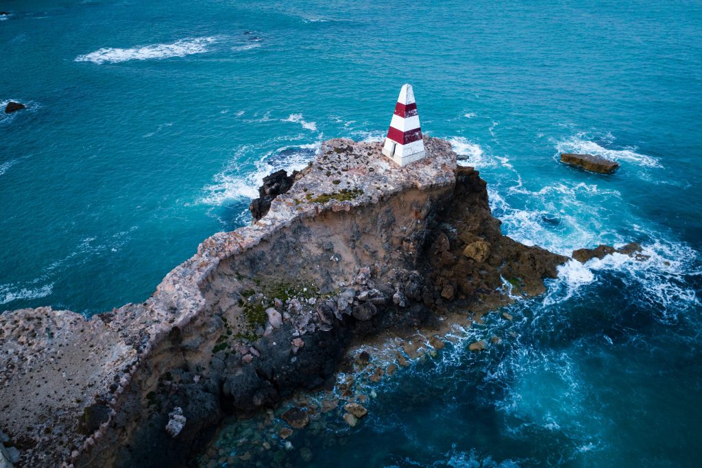 Aerial view of a striped lighthouse on a rugged coastal promontory surrounded by blue sea.
