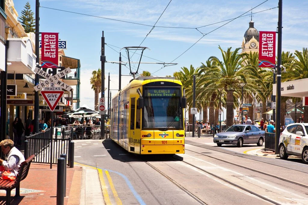 A yellow tram travels on a street in Glenelg, lined with palm trees, shops, and pedestrians. A "Jetty Road Glenelg" sign and a triangular traffic sign are visible.