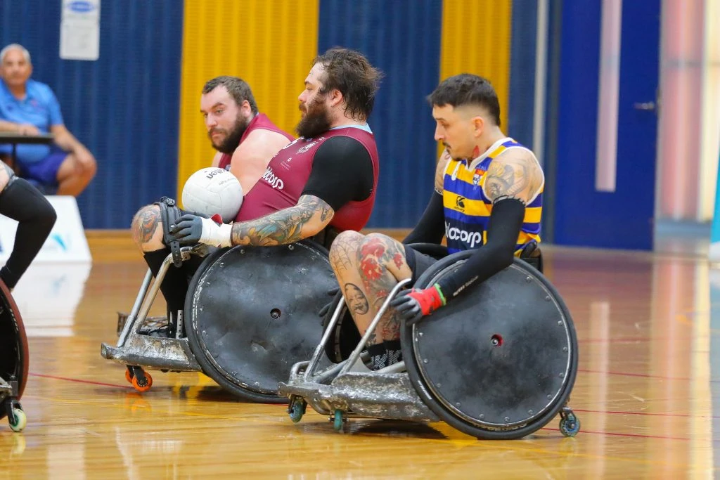 Three athletes in wheelchairs compete in a rugby game. Two wear maroon jerseys, and one wears a blue and yellow striped jersey. One athlete holds a white ball. A person observes in the background.