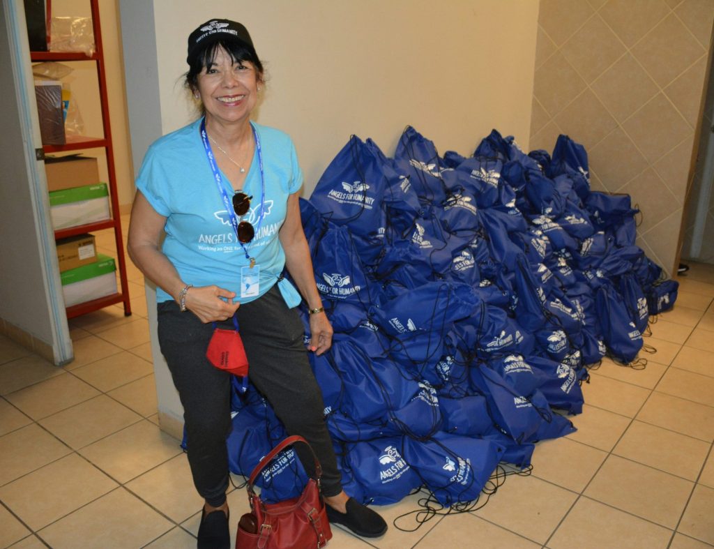 A woman in casual attire stands next to a large pile of blue drawstring bags in a tiled room. She wears a black cap and holds a red bag while smiling at the camera.