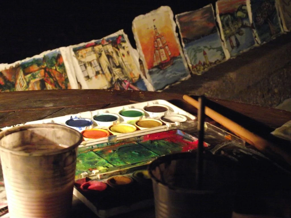 A table with a paint palette, brushes, and cups. Several watercolor paintings are displayed in the background, depicting various scenes.