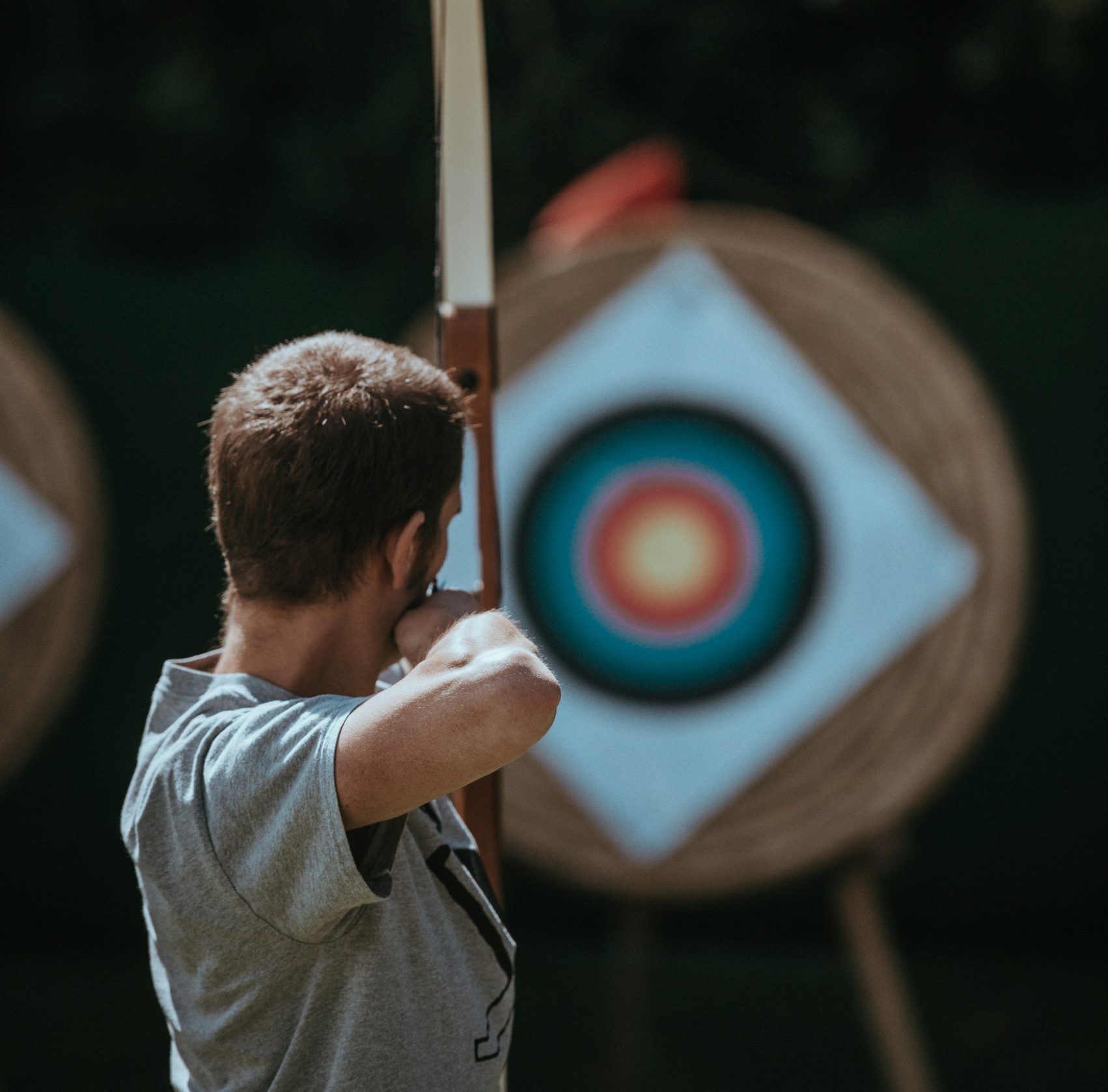 A person in a grey shirt aims a bow and arrow at a bullseye target during an archery practice session.