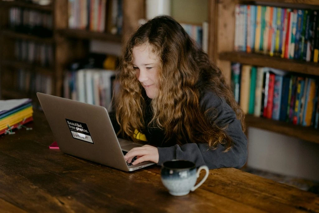 A child with long curly hair uses a laptop at a wooden table with a bookshelf in the background and a coffee mug beside the laptop.