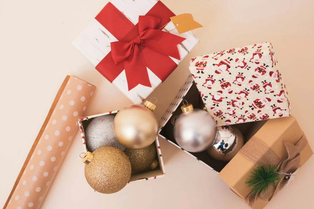 Open gift boxes with red and white wrapping paper, containing gold and silver Christmas ornaments, beside rolled polka dot wrapping paper.