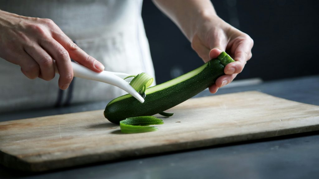 Hands using a white peeler to slice a green zucchini on a wooden cutting board.