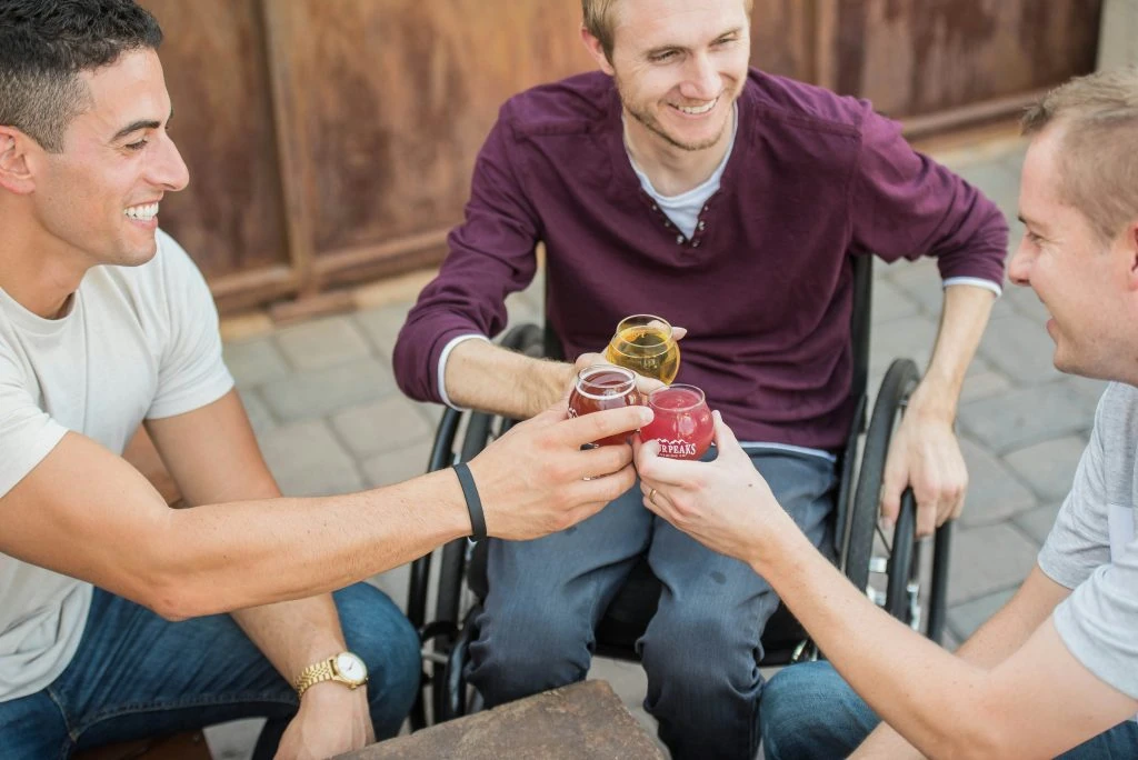 Three men, one in a wheelchair, are sitting outdoors and clinking small glasses together, smiling and enjoying a drink.