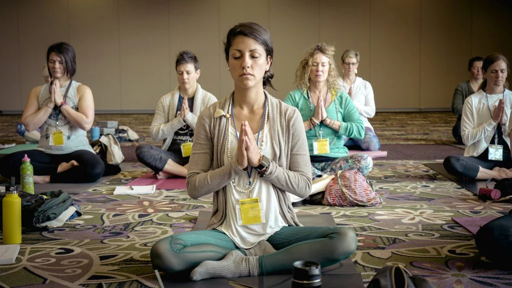 A group of people sitting cross-legged on yoga mats with eyes closed, meditating indoors.