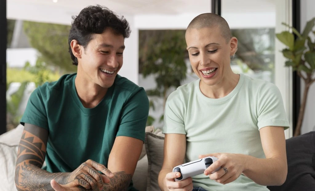 Two people sitting on a couch, smiling and interacting with a video game controller in a bright, modern living room.