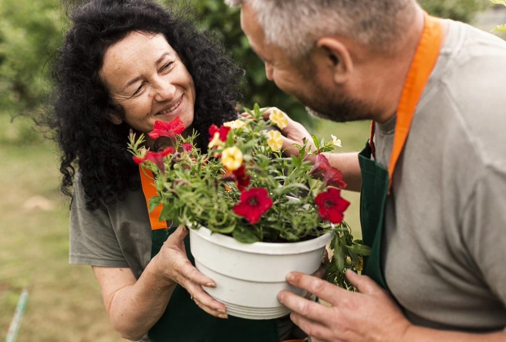 Two people in aprons smiling at each other while holding a pot with red and yellow flowers outdoors.