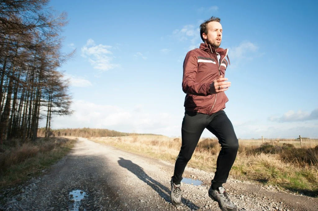 A person in a maroon jacket and black pants runs on a gravel path in a rural, sunlit area.