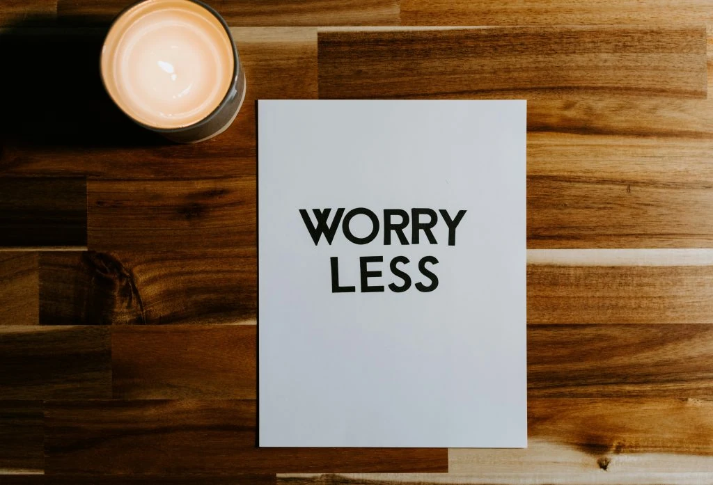 A lit candle and a piece of paper with the words "WORRY LESS" printed on it sit on a wooden surface.