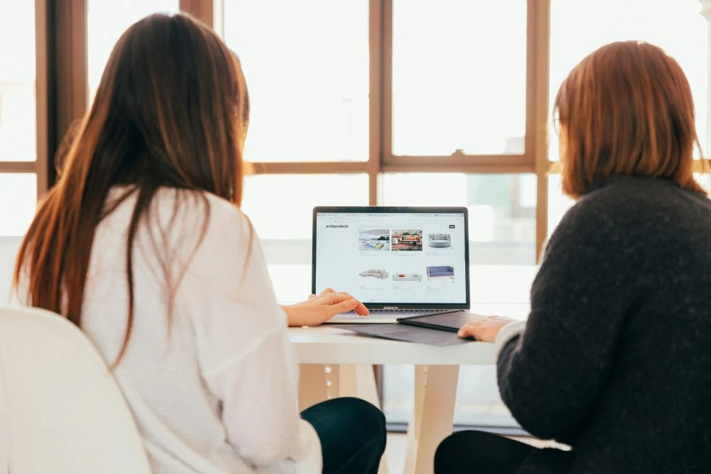 Two people sit at a table, looking at a laptop screen displaying a website with various images and options.