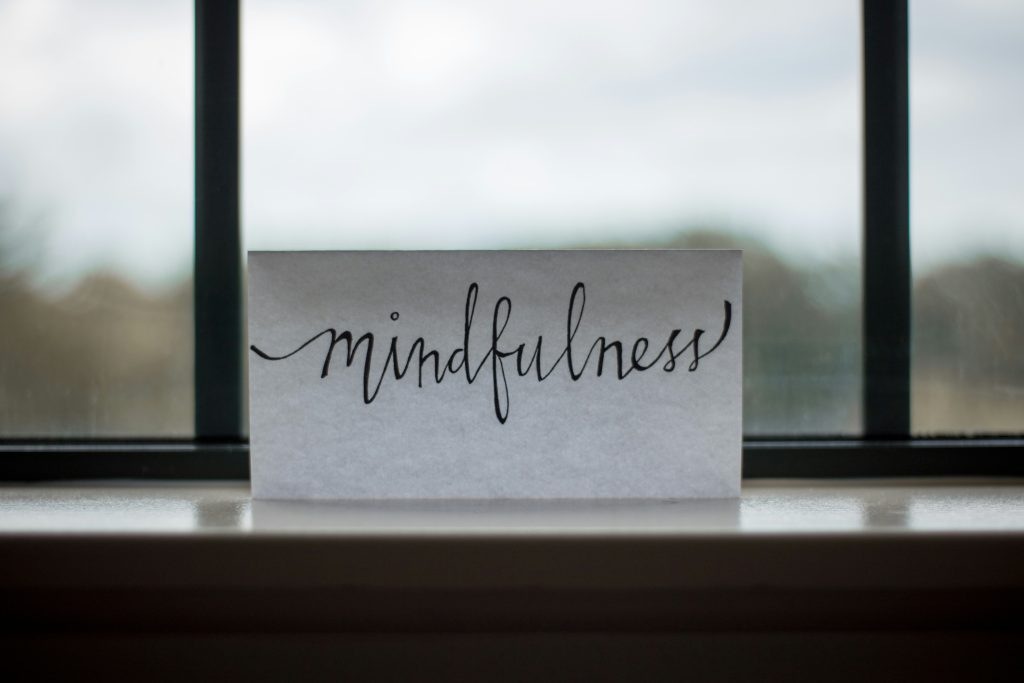 A white card with the word "mindfulness" written in cursive is placed on a windowsill with an out-of-focus background.