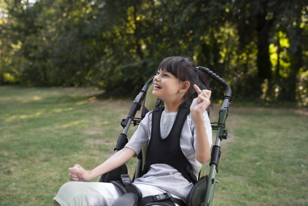 A young girl in a wheelchair smiles and gestures while sitting outside on grassy terrain, surrounded by trees.