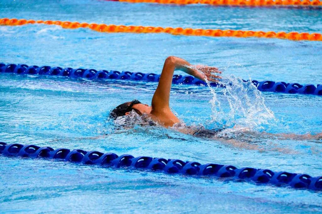 Swimmer wearing goggles and a swim cap performing the backstroke in a swimming pool lane marked by blue and orange lane dividers.