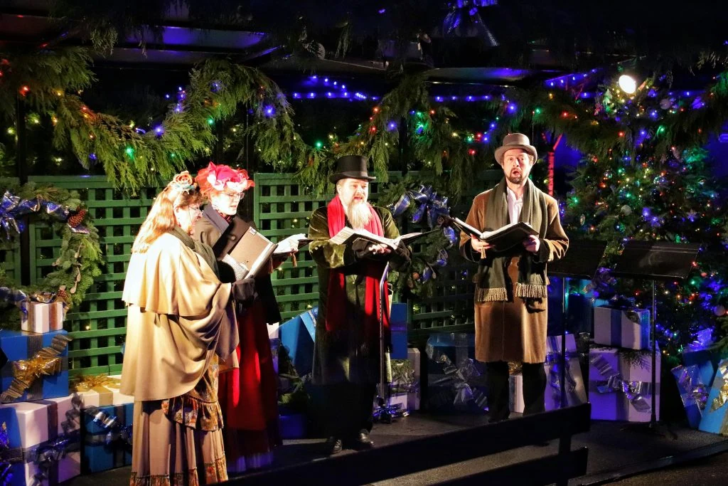 Four people in period costumes sing carols under festive lights, surrounded by decorated trees and wrapped presents.