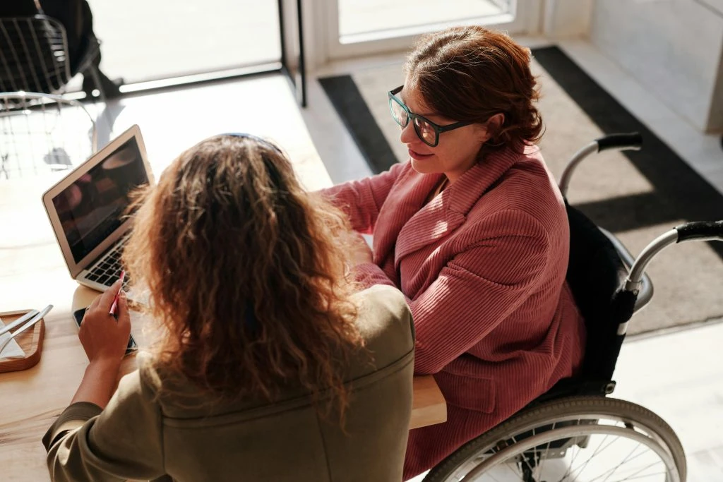 Two people at a table with a laptop; one wearing a pink jacket and glasses, uses a wheelchair. They are in conversation in a naturally lit room.