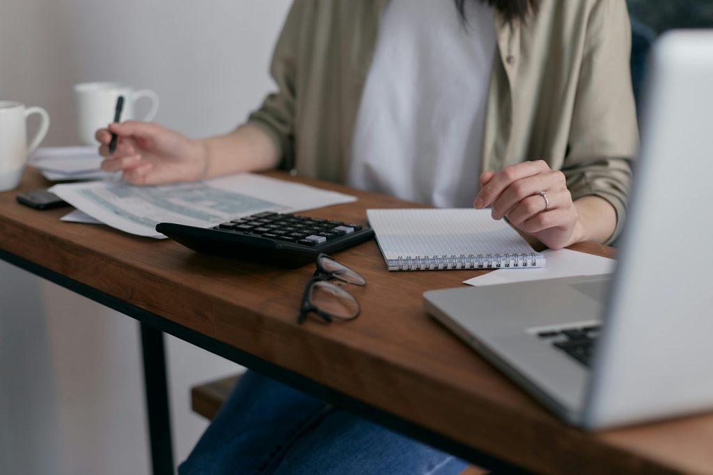 Person sitting at a desk with a calculator, papers, a notebook, and a laptop, holding a pen. Glasses rest on the desk.