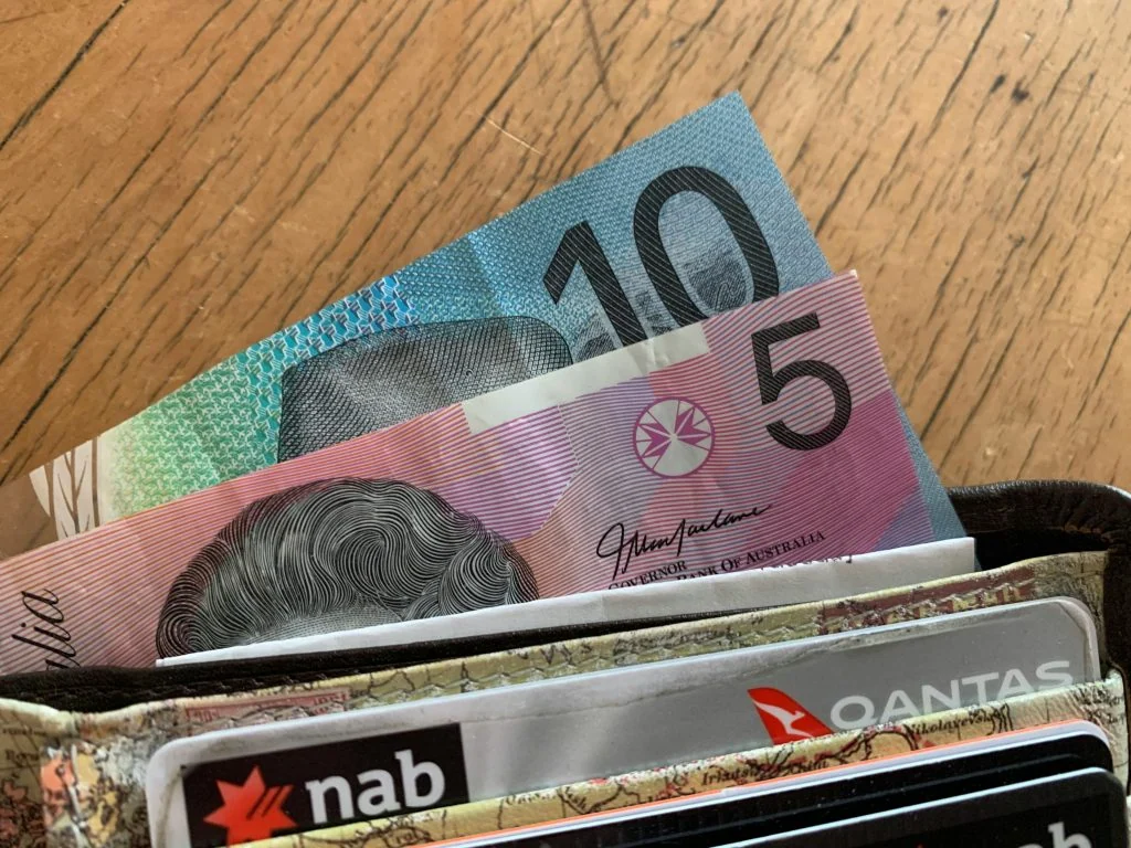 A wallet with Australian 10 and 5 dollar notes and various bank cards, including one from NAB and a Qantas card, on a wooden surface.