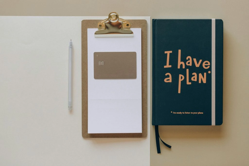 A pen, a clipboard holding a card, and a dark green notebook with "I have a plan!" on the cover are neatly arranged on a beige surface.