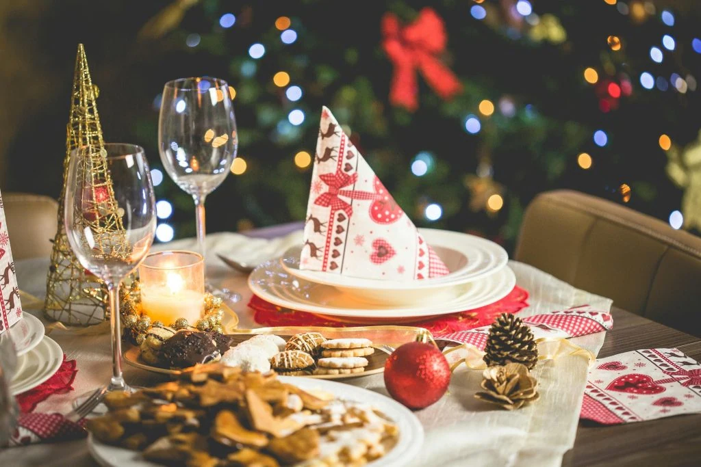 Festive dining table set with plates, glasses, napkins, and baked goods. Christmas tree with lights in the background.