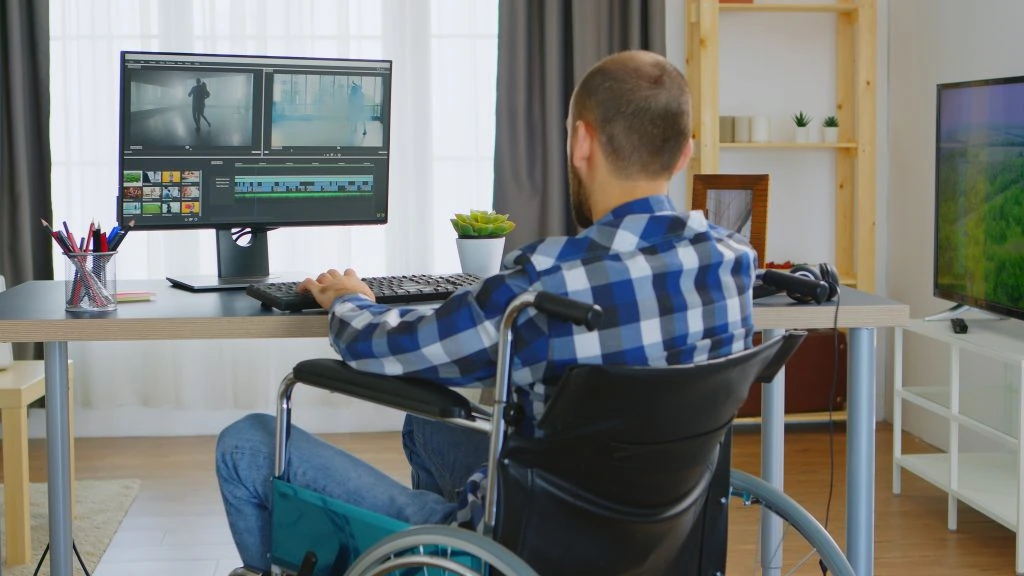 A person in a wheelchair sits at a desk using a computer to edit video footage in a modern, well-lit room.