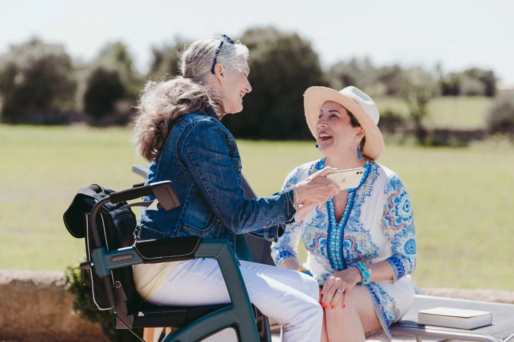 Two women are seated outdoors, chatting and smiling. One woman uses a mobility aid and holds a smartphone. The background shows greenery and a sunny sky.