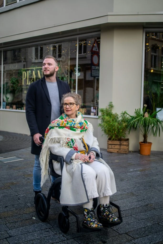 A man pushes a woman in a wheelchair on a city street. The woman is wearing a colorful scarf and patterned shoes, while the man is dressed in a dark coat and jeans.