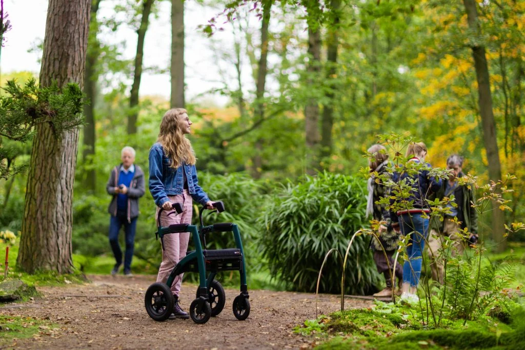 A woman with a walker stands on a forest path, looking up. Several other people walk in the background.