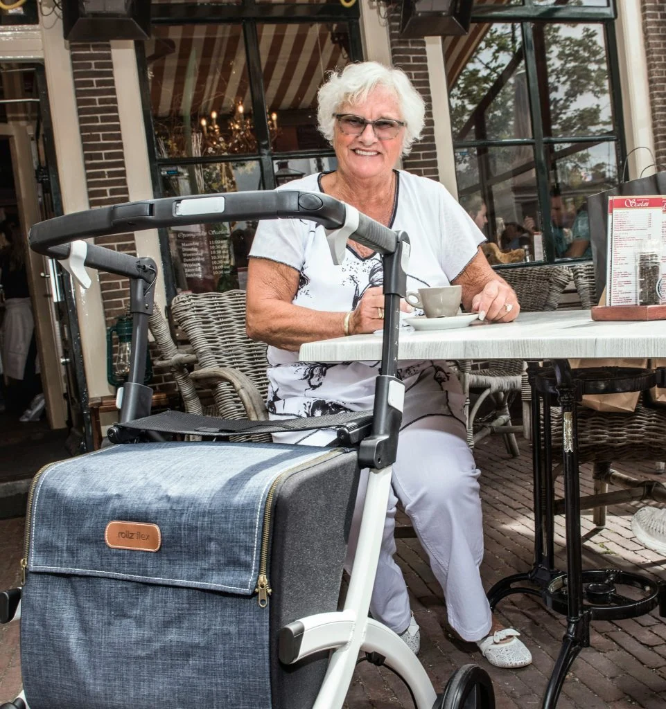 Elderly woman sitting at an outdoor café table, smiling, with a walker in front of her.