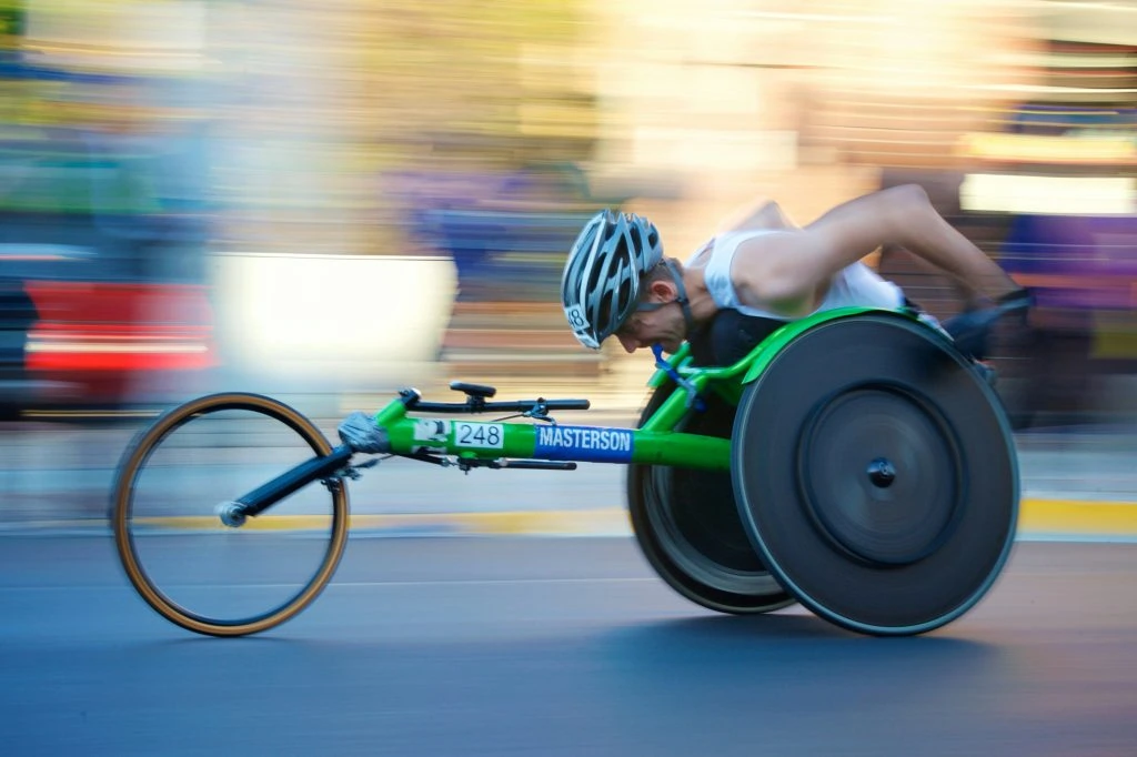 A person competes in a racing wheelchair, wearing a helmet and a tank top, with blurred background indicating motion and speed.