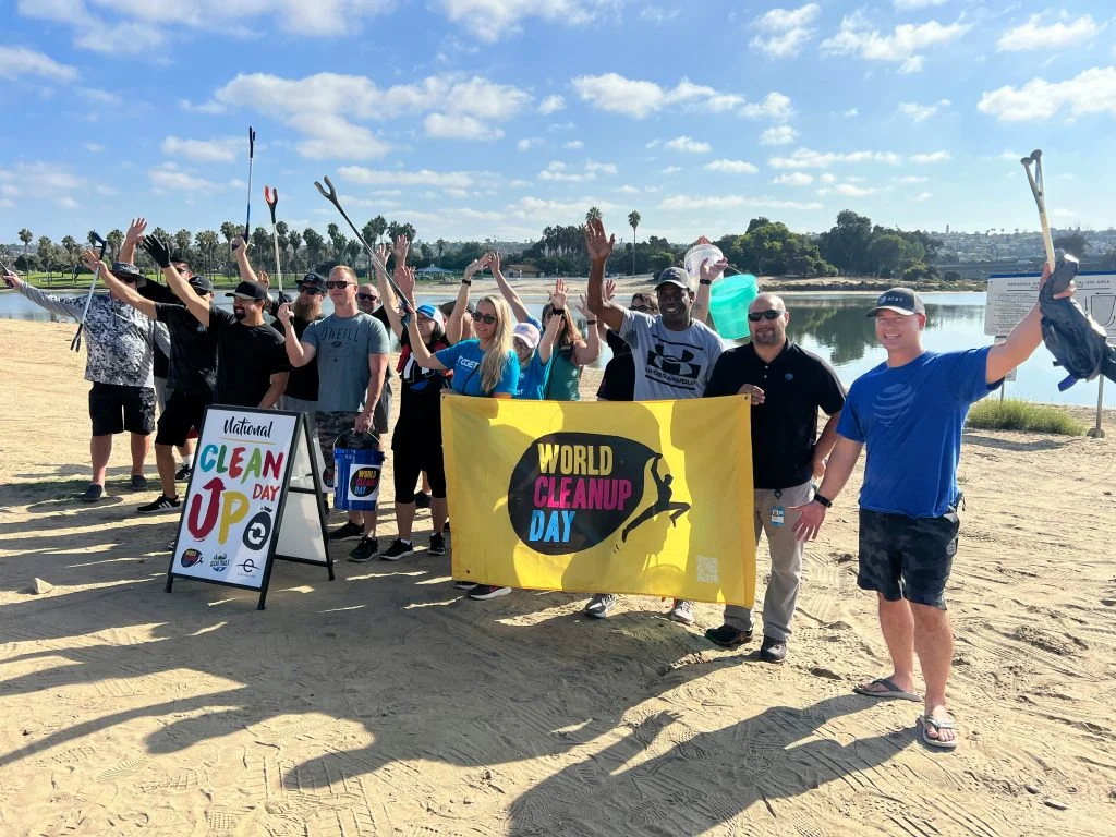 A group of people on a beach hold up litter pickers and a "World Cleanup Day" banner, with a sign and a calm body of water in the background.