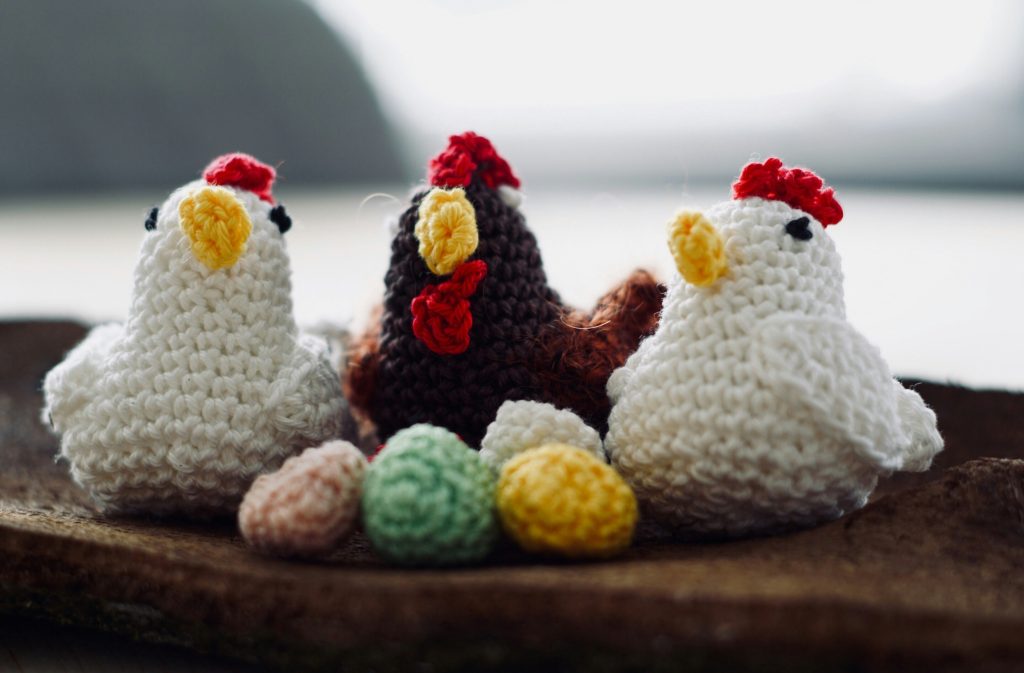 Three crochet chickens with colorful eggs on a wooden surface.