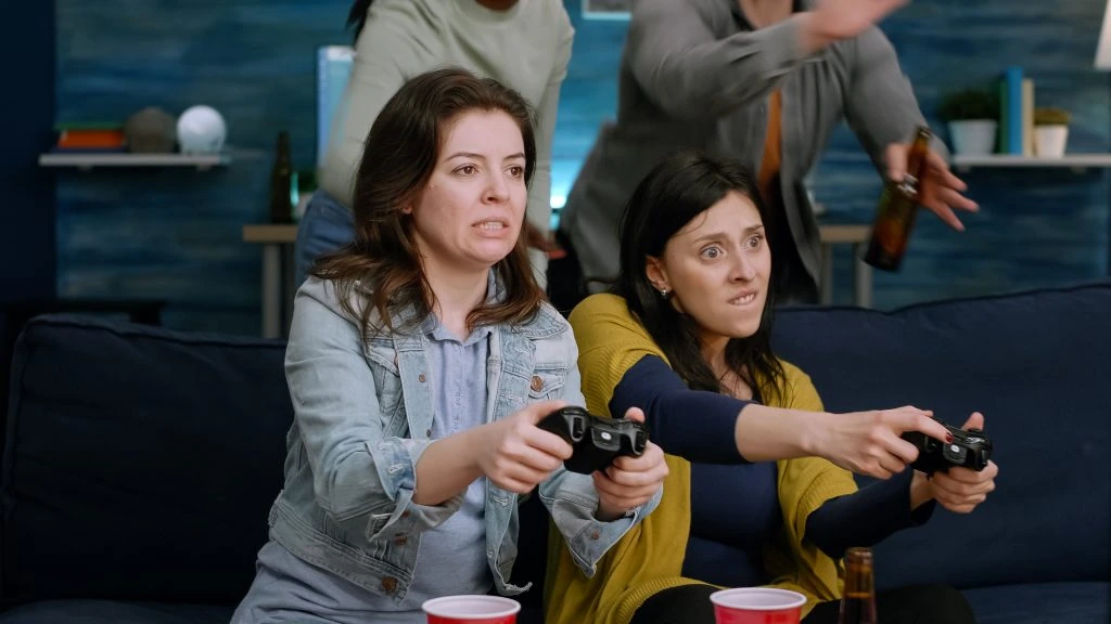 Two women sit on a couch holding game controllers, focused on a video game, with drinks and people in the background.