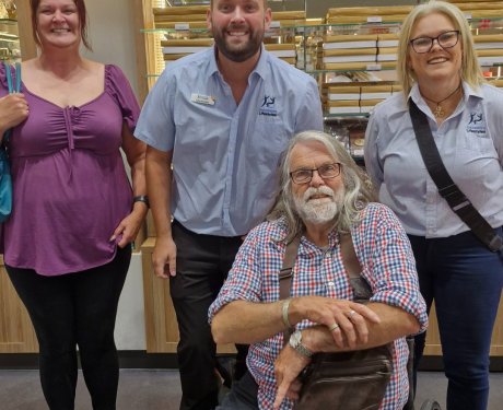 Four people posing indoors, with two standing on each side of a man sitting in a wheelchair. They are in a room with shelves in the background.