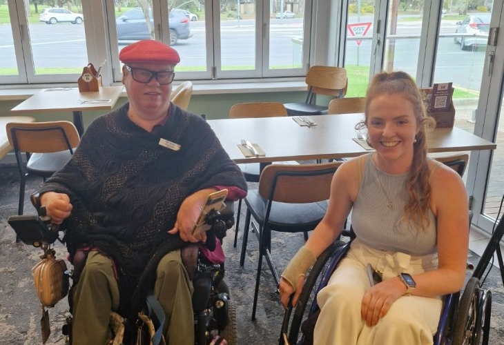 Two people in wheelchairs are sitting next to each other in a dining area. One person is wearing a red hat, glasses, and a dark poncho. The other person is wearing a light-colored sleeveless top and white pants.