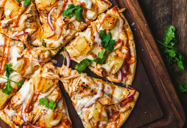 Pizza topped with pineapple, chicken, red onion, and cilantro on a wooden board, with cilantro leaves scattered around.