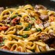 A close-up of a pan filled with cooked fettuccine pasta mixed with pieces of beef, sautéed spinach, cherry tomatoes, and mushrooms.