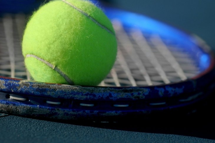 A close-up of a tennis ball resting on a blue tennis racket, with the focus on the ball and stringed surface of the racket.