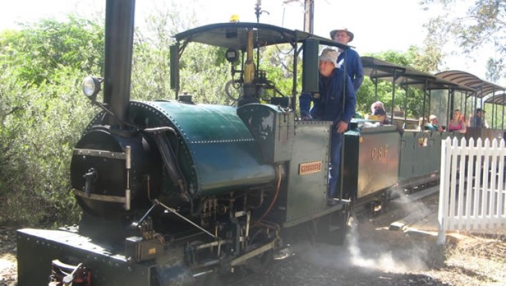 Front view of a mini steam train. There are two older men dressed in blue overalls driving the engine and the carriages with people in them are following.