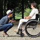 A woman in a hat squats and converses with a smiling woman in a wheelchair on a park pathway surrounded by trees.