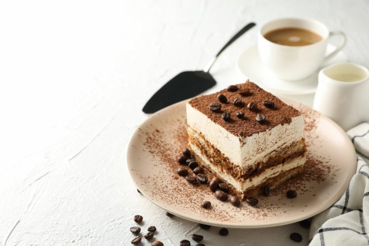 A large square piece of tiramisu cake sprinkled with coffee beans. Behind the plate of cake is a white coffee mug