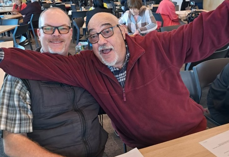 Two men are seated at a table in a crowded dining area. One man is smiling, while the other is extending his arms joyfully. Papers are on the table in front of them.