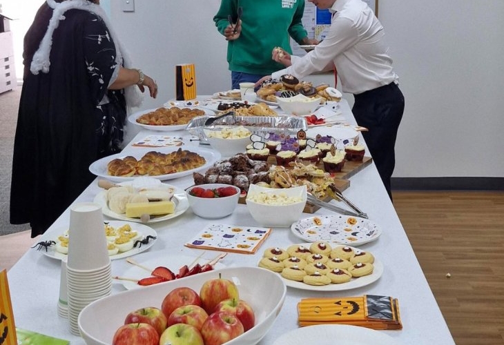 People in costumes stand by a table with Halloween-themed food, including apples, cookies, and pastries.