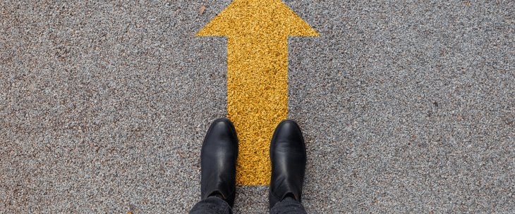 A yellow arrow painted on concrete with a man's boots standing next to it